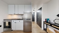 This thoughtfully designed studio apartment at 111 Porter Avenue in Buffalo, NY, offers a clean, modern aesthetic throughout its single-floor open layout. Light-toned hardwood flooring runs continuously through the entire space, lending warmth and visual cohesion to every area.

The kitchen is well-appointed with crisp white cabinetry featuring matte black hardware, dark countertops, a white subway tile backsplash, and stainless steel appliances including a top-freezer refrigerator and a freestanding range. A peninsula with additional counter space provides a practical divide between the kitchen and the living area.

The entry corridor features dark charcoal doors — including closet and unit entry — accented by a stylish console table that creates a welcoming foyer moment. Recessed and flush-mount ceiling lighting keeps the space bright and functional.

The sleeping and living area is open and airy, anchored by a large window fitted with horizontal blinds that invites natural light. The layout comfortably accommodates a full-sized bed, a lounge chair, and additional seating, making it a versatile and livable space. Neutral white walls throughout provide a clean backdrop that complements the warm, modern furnishings and décor.