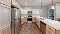 This single-floor residence is designed with a clean, contemporary aesthetic carried consistently throughout every room. The kitchen serves as a functional centerpiece, featuring an L-shaped layout with a combination of light wood and white shaker-style cabinetry, white quartz countertops, stainless steel appliances including a refrigerator, range, microwave, and dishwasher, and a peninsula with a pendant light overhead. Wood-look plank flooring extends seamlessly from the kitchen into the open living area, which benefits from a large sliding glass door fitted with vertical blinds, drawing in natural light and providing outdoor access.

The bedrooms maintain the same neutral gray palette and wood-look flooring, each equipped with windows fitted with blinds. One bedroom features a ceiling fan, while another includes closet access. The bathroom offers a single-sink vanity with light wood cabinetry, an integrated mirror, built-in shelving, and a full bathtub with a tub/shower combination. A dedicated laundry area with a washer and dryer is also present. Recessed lighting and louvered closet doors contribute to the space's polished, modern character throughout.