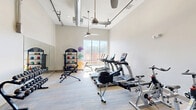 Located at 5437 Bowman Rd in Macon, Georgia, this dedicated fitness center at Lofts at Bass offers a well-appointed exercise space designed to support a comprehensive workout experience. The room features soaring ceilings with exposed industrial-style ductwork and ceiling fans that promote airflow and contribute to a modern, open aesthetic. Large windows along one wall flood the space with natural light and provide views of the surrounding greenery, creating an energizing environment.

The fitness center is outfitted with a diverse selection of equipment to accommodate cardio and strength training needs. Cardio equipment includes treadmills, rowing machines, and stationary spin bikes. A multi-station cable machine supports resistance and strength training, while a well-stocked free weight rack offers a full range of dumbbells. Organized equipment racks hold medicine balls, stability balls, kettlebells, and resistance bands, providing options for dynamic and functional fitness routines.

A wall-mounted flat-screen television enhances the workout experience, and a water cooler station keeps users hydrated. Full-length mirrors are positioned along one wall to assist with form and technique. The flooring features durable, low-pile carpet tile suited for a high-traffic fitness environment. Glass-panel entry doors and clear sightlines contribute to the room's bright, accessible feel. Overall, this fitness center delivers a fully functional and thoughtfully designed space for residents seeking convenient on-site exercise amenities.