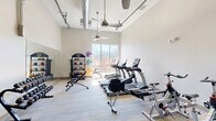 Located at 5437 Bowman Rd in Macon, Georgia, this dedicated fitness center at Lofts at Bass offers a well-appointed exercise space designed to support a comprehensive workout experience. The room features soaring ceilings with exposed industrial-style ductwork and ceiling fans that promote airflow and contribute to a modern, open aesthetic. Large windows along one wall flood the space with natural light and provide views of the surrounding greenery, creating an energizing environment.

The fitness center is outfitted with a diverse selection of equipment to accommodate cardio and strength training needs. Cardio equipment includes treadmills, rowing machines, and stationary spin bikes. A multi-station cable machine supports resistance and strength training, while a well-stocked free weight rack offers a full range of dumbbells. Organized equipment racks hold medicine balls, stability balls, kettlebells, and resistance bands, providing options for dynamic and functional fitness routines.

A wall-mounted flat-screen television enhances the workout experience, and a water cooler station keeps users hydrated. Full-length mirrors are positioned along one wall to assist with form and technique. The flooring features durable, low-pile carpet tile suited for a high-traffic fitness environment. Glass-panel entry doors and clear sightlines contribute to the room's bright, accessible feel. Overall, this fitness center delivers a fully functional and thoughtfully designed space for residents seeking convenient on-site exercise amenities.