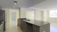This single-floor space showcases a contemporary design with a clean, minimalist aesthetic throughout. The kitchen serves as a central feature, anchored by a substantial island that offers generous countertop space finished in a dark stone-toned surface. The island is equipped with an integrated sink and a stainless steel dishwasher, complemented by medium-toned wood-grain cabinetry with sleek bar-pull hardware beneath. Additional counter space lines the perimeter, providing practical workspace for cooking and preparation. The cabinetry carries a consistent warm wood-grain finish that contrasts effectively against the light-toned tile flooring, which extends throughout the open area. Walls are finished in a crisp off-white, contributing to a bright and airy atmosphere. Recessed lighting is embedded in the ceiling, ensuring even illumination across the space. A frosted-glass door is visible in the background, allowing diffused natural light to filter in while maintaining privacy. Ceiling-mounted HVAC vents suggest a well-integrated climate control system. The open layout creates a fluid, unobstructed flow between areas, making the space feel both functional and inviting.