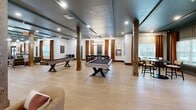 Located at 13100 Margie Ln in Raleigh, NC, this single-story community clubhouse offers a generously sized, thoughtfully designed interior spread across one open floor. The space is defined by a cohesive modern industrial aesthetic, featuring wide-plank light hardwood flooring, dark painted exposed beam ceilings with teal accents, and sturdy wood-wrapped structural columns that anchor each zone with warmth and character.

The leasing and reception area greets visitors with a sleek front desk, built-in shelving displays, and lush greenery climbing the central columns, creating a welcoming first impression. Adjacent to this is a spacious lounge area furnished with plush gray sofas, velvet accent chairs, and a mix of pendant and globe lighting suspended from the dramatic ceiling — ideal for relaxed gatherings.

A dedicated games room provides ample space for a full-size billiards table and shuffleboard, flanked by windows dressed in warm burnt-orange drapes that fill the area with natural light. Additional seating alcoves and casual lounge nooks are scattered throughout, offering intimate settings for socializing or working. The space also includes restrooms and private office areas. Throughout, curated artwork, decorative shelving, and layered lighting combine to create an inviting, upscale communal atmosphere.