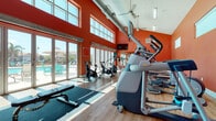 Located at 5900 Wilkinson Rd in Sarasota, Florida, this single-floor fitness center is designed to energize and inspire with its bold, vibrant interior. The space is immediately distinguished by its soaring vaulted ceilings and an expansive bank of floor-to-ceiling sliding glass doors and clerestory windows, flooding the interior with abundant natural light and offering direct views of the outdoor pool area complete with lounge chairs and umbrellas.

The warm terracotta-orange walls lend a dynamic, motivating atmosphere throughout the space, complemented by durable wood-look flooring. Ceiling fans are positioned overhead to maintain comfortable airflow. The fitness floor is well-equipped with a comprehensive selection of cardio machines, including treadmills, ellipticals, stair climbers, and stationary and spin bikes. A dedicated strength training area features a full rack of dumbbells, kettlebells, and stability balls, with wall-mounted mirrors enhancing the sense of space and supporting proper form during workouts.

A mounted flat-screen television is visible from the main exercise area, and an air conditioning unit ensures year-round comfort. Additional amenities include a water bottle refill station and drinking fountain, a small refreshment and information table, and a restroom accessible from within the facility. The seamless connection between the bright, open interior and the resort-style pool area outside makes this fitness center a standout feature of the property.