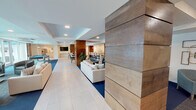 This single-floor amenity space is thoughtfully designed to accommodate a variety of social and leisure activities. The layout flows seamlessly from one area to the next, beginning with an open lounge zone anchored by neutral-toned sofas, accent chairs, and light tile flooring. Substantial wood-clad columns add a warm architectural contrast against the crisp white ceilings with recessed lighting throughout.

A generously sized bar counter, topped with a striking granite surface featuring bold gold and black veining, serves as a central gathering point. Bar-height seating lines the counter, which faces a wall-mounted flat-screen TV. Adjacent to the bar, a dining and café-style seating area provides additional space for socializing, furnished with high-top tables and chairs arranged beneath tray ceilings with subtle blue accents.

The game room area features a full-size pool table set on rich blue carpet, while oversized floor-to-ceiling windows flood the space with natural light and provide direct views of the outdoor pool, putting green, and surrounding palm-lined terrace. A separate, private meeting room offers a quieter retreat, with wood-look flooring, panel lighting, and a conference-style table for small gatherings. Together, these areas create a well-rounded, amenity-rich environment.