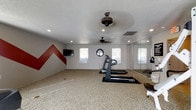 This single-floor fitness space is thoughtfully designed to serve as a fully functional workout room. The room features a speckled, durable floor covering well-suited for exercise activities, providing both comfort and practicality underfoot. A treadmill and stair-climber are positioned centrally, offering cardio options within a spacious, open layout. A full-length mirrored wall along one side enhances the sense of space while allowing users to monitor their form during workouts. The room is brightened by multiple windows that bring in natural light, creating an energizing atmosphere. Two ceiling fans ensure proper air circulation throughout the space, keeping the environment comfortable during intense sessions. Recessed lighting supplements the natural light for well-distributed illumination at any time of day. A wall-mounted flat-screen television adds an entertainment element to workouts. A bold red chevron accent painted on one wall adds a dynamic, motivational visual focal point. The textured ceiling completes the room's character. Overall, this fitness room balances functionality with design, offering a well-equipped, purposeful environment for daily exercise routines.