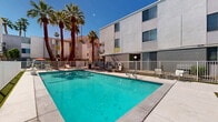 Located in the heart of Palm Springs, California, this generously sized outdoor pool and courtyard space offers a quintessential desert resort experience. The centerpiece is a large, rectangular swimming pool with clear turquoise water, framed by a wide concrete pool deck enclosed within a clean white metal fence. The surrounding multi-story buildings feature a modern, mid-century-influenced architectural style with smooth stucco exteriors, corrugated metal cladding accents, and vibrant chartreuse yellow entry doors that add a bold pop of color to the neutral palette.

Towering palm trees punctuate the skyline, lending the courtyard a lush, tropical character while framing dramatic views of the rugged San Jacinto Mountains rising in the background. The pool deck is well-appointed with lounge chairs, chaise lounges, and shaded seating areas complete with green market umbrellas, providing ample space for relaxation under the abundant Southern California sunshine. Manicured turf strips line the perimeter pathway, adding a touch of greenery against the desert landscape.

The open-air layout of the courtyard is bright and airy, benefiting from exceptional natural light year-round. Accessed via gated entry points, the space is both private and inviting, capturing the relaxed yet stylish spirit that defines Palm Springs living.