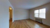 This single-floor space presents a bright and straightforward layout centered around a generously sized main room. The room features warm-toned hardwood floors throughout, lending a classic and inviting character to the interior. Crisp white walls and a smooth white ceiling create a clean, neutral backdrop well-suited to a variety of uses and décor styles. A double-hung window on one wall allows natural light to filter into the space, complemented by a cast-iron radiator positioned beneath it — a detail indicative of traditional construction and a reliable heating method. An elegantly curved archway on the opposite side of the room provides a graceful transition into an adjoining area, adding a touch of architectural charm that distinguishes the space from more modern, utilitarian designs. The archway hints at a connected hallway or additional room beyond. Overall, the space maintains a simple, uncluttered ambiance with its unfurnished state making the structural and design elements especially apparent. The combination of hardwood flooring, arched doorway, and radiator heating reflects the character of classic residential construction.