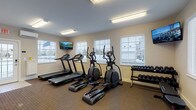 Located at 7086 Crown Lake Dr in Reynoldsburg, Ohio, this single-floor fitness center is a well-equipped exercise facility designed to support a variety of workout routines. The space is thoughtfully laid out with a carpeted workout area and tile flooring near the entry, creating clear zones for different activities.

The main exercise area is outfitted with multiple treadmills and elliptical cross-trainers, all from the TM6000 and FL6000 series, providing ample cardio options for users. A dumbbell rack stocked with a range of free weights offers strength training capabilities, while a weight bench and a posted stretching guide chart support resistance and flexibility work. Wall-mounted flat-screen TVs are positioned throughout the room, allowing users to stay entertained during their workouts.

Natural light fills the space through several windows fitted with white horizontal blinds, offering views of the surrounding exterior. The room is climate-controlled via a wall-mounted mini-split HVAC unit, ensuring comfort year-round. A dedicated entryway with a glass-panel door, coat hooks, a water fountain, fire safety equipment, and a waste bin rounds out the practical amenities. The overall ambiance is clean, functional, and well-maintained, making it a convenient fitness resource for the Crown Lake community.