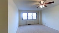 This single-floor space presents a clean and straightforward layout designed for comfortable living. The main room is finished with wall-to-wall carpeting in a neutral tone, contributing to a calm and understated ambiance throughout. A triple-panel window serves as the focal point of the primary wall, allowing an abundance of natural light to flood the interior and creating an open, airy feel. Beneath the window sits a radiator heating unit, providing an efficient and classic approach to climate control. The ceiling is fitted with a multi-blade ceiling fan with an integrated light fixture, offering both ventilation and supplemental lighting to the room. The walls are finished in a crisp, light neutral tone, enhancing the brightness of the space and providing a versatile backdrop. The overall design is practical and uncluttered, with clean lines and a simple palette that makes the space feel welcoming and easy to personalize. The combination of natural light, soft carpeting, and functional fixtures creates a livable environment well-suited for everyday use.