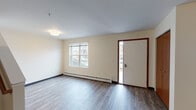 This two-story unit is designed with a practical and clean aesthetic throughout. The main level features an open entryway and living area with durable hardwood-style flooring, cream-toned walls, and wood-trimmed doors that add warmth to the interior. A staircase with a handrail leads to the upper floor, and baseboard heating units are present throughout for consistent climate control. A wall-mounted mini-split unit in the kitchen area provides additional climate management.

The kitchen is well-appointed with rich dark wood cabinetry extending to the ceiling, light countertops, a double stainless steel sink, and a full suite of black appliances including a refrigerator, electric range, microwave, and dishwasher. Track lighting illuminates the space effectively.

The bathrooms feature hardwood-style flooring, dark vanity cabinetry, white countertops, and a tub-shower combination. One bathroom includes a stacked washer and dryer unit conveniently built into an alcove. A half bath provides an additional toilet and storage area. Upstairs, carpeted bedrooms offer a softer feel, with windows allowing natural light and exterior views.
