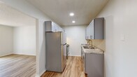 This single-floor unit at 1717 S Luster Ave in Springfield, Missouri offers a well-organized layout connecting living, dining, kitchen, and bedroom areas through a central hallway. The kitchen is designed as a galley-style space, featuring gray shaker cabinetry with brushed nickel hardware, granite-patterned countertops, a double-basin sink, and stainless steel appliances including a refrigerator and range. Recessed lighting illuminates the kitchen, complementing the light wood-tone luxury vinyl plank flooring that runs throughout the main living areas and hallway. The living room is bright and open, with a sliding glass door fitted with vertical blinds that invites natural light and provides access to an outdoor area. A textured ceiling adds architectural character to the main living space. The hallway connects the living areas to the bedrooms, which feature plush carpeting, ceiling fans, and natural light from windows with blinds. At least one bedroom includes a closet for dedicated storage. The bathroom is equipped with a clean, white tub and shower surround with brushed nickel fixtures. Throughout, neutral white walls and consistent flooring create a cohesive, move-in-ready environment.