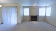 This single-floor residence offers a comfortable and functional living space with a clean, neutral aesthetic throughout. The main living area is generously sized, featuring wall-to-wall carpeting that adds warmth underfoot and a cohesive, inviting feel to the room. A centrally positioned fireplace serves as the focal point of the space, framed by a sleek tile surround and hearth that provides both visual interest and a cozy gathering spot. The walls are finished in a soft, neutral tone that complements the light carpeting and contributes to the overall bright, airy ambiance. Natural light flows into the living area through multiple windows fitted with white horizontal blinds, allowing for adjustable illumination throughout the day. A set of vertical blinds covers what appears to be a sliding glass door, offering convenient access to an outdoor area while maintaining privacy. A flush-mount ceiling light fixture provides additional overhead illumination. The layout is open and unobstructed, creating a versatile space well-suited for a variety of furniture arrangements. The combination of the fireplace, ample natural light, and neutral finishes gives this residence a welcoming and move-in-ready character.