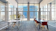 This contemporary collaborative lounge and workspace is designed to inspire productivity and creativity through its striking architectural features and thoughtful layout. The space is defined by soaring double-height ceilings finished in a bold dark blue-gray, creating a dramatic contrast against the crisp white walls below. An expansive wall of floor-to-ceiling windows and glass doors floods the interior with abundant natural light and provides sweeping views of the surrounding urban skyline, giving the space an open, airy feel.

The main area features a mix of flexible furnishings, including marble-topped tables paired with modern chairs in warm terracotta and light mint tones, accommodating both collaborative group work and individual use. A patterned blue-gray carpet grounds the space and adds texture underfoot. Edison-style pendant lights hang from the high ceiling, contributing a warm, industrial-chic aesthetic.

A glass-partitioned private meeting room sits along one side of the space, enclosed by sleek steel-framed glass panels and accessed through a glass door, offering a quiet enclave for focused meetings or calls. Adjacent to this room, a built-in workstation with a wood-and-stone counter provides a dedicated spot for printing and administrative tasks. The overall ambiance is modern, professional, and welcoming, well-suited for coworking, meetings, and community gatherings.