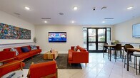 The Julien Clubroom at 2142 Commonwealth Ave in Charlotte, NC is a well-appointed community amenity space designed for socializing and relaxation. The single-floor layout is finished with large-format tile flooring throughout, recessed lighting, and crisp white walls that create a bright, welcoming atmosphere.

The main lounge area is anchored by bold mid-century modern furniture, including vibrant red sofas and accent chairs arranged around a slatted wood coffee table, all centered beneath a large wall-mounted flat-screen television. A colorful large-scale abstract painting adds a lively artistic focal point to the room, complemented by decorative ceramic vases on a built-in ledge shelf.

Adjacent to the lounge is a dining area featuring high-top wood tables with modern chairs, a second wall-mounted television, and floor-to-ceiling glass doors that open directly to an outdoor patio, flooding the space with natural light and providing seamless indoor-outdoor connectivity.

The kitchenette is equipped with granite countertops, white cabinetry, a coffee and beverage station, a microwave, a wet bar sink, and an under-counter beverage refrigerator — making it well-suited for hosting gatherings. Warm wood-panel wall accents in this area add texture and visual interest.

The space is accessed through a set of elegant glass-paneled double doors marked with The Julien branding, reinforcing the polished, community-centered identity of this clubroom.