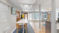 Located at 7301 Bristol Village Dr in Bloomington, MN, this single-floor commercial space offers a thoughtfully designed layout that balances professional functionality with an inviting atmosphere. The interior is organized into distinct zones that flow seamlessly into one another, supported by warm hardwood flooring and neutral gray walls throughout.

The reception area serves as the focal point upon entry, anchored by a curved, textured front desk finished in a woven wood-tone material. Pendant and flush-mount lighting illuminate the space warmly, while rich brown leather seating and a colorful teal-patterned area rug create a comfortable waiting environment. A wall-mounted flat-screen TV adds a modern, informative touch.

A central corridor features deep blue carpet and a raised communal bar-height table with wooden stools, functioning as a collaborative or casual meeting area. Warm-toned wood-framed glass doors lead to an adjacent room visible through a large interior glass wall, offering a glimpse of a secondary lounge space with a fireplace.

A standout architectural feature is the coffered ceiling with integrated skylights, which floods the main gathering area with natural light. Large bay and picture windows along the perimeter provide expansive outdoor views, further enhancing the bright, open ambiance of the space.