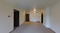 This single-floor residential space offers a straightforward and functional layout designed for comfortable everyday living. The main living area is generously sized, finished with wall-to-wall carpeting that adds warmth and softness underfoot. A ceiling fan with an integrated light fixture is centrally mounted, providing both air circulation and ambient lighting throughout the room.

The space features neutral white walls and dark-trimmed doorways that create a clean visual contrast throughout the interior. A solid wood entry door is visible at the far end of the living area, serving as the main point of entry. Additional doorways branch off to the left and right, suggesting access to further rooms such as bedrooms, a kitchen, or storage areas.

A floor-level vent indicates a forced-air heating and cooling system, ensuring year-round climate control. The layout is open and unobstructed, allowing for flexible furniture arrangement to suit a variety of needs. The overall ambiance is clean, understated, and move-in ready, with a no-frills design that prioritizes practicality and livability. The consistent use of neutral tones throughout creates a calm, cohesive atmosphere that serves as a versatile backdrop for personalization.