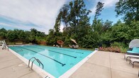 This community pool facility is built around a generously sized outdoor lap pool with clear, turquoise water and marked lanes, surrounded by a wide concrete deck outfitted with rows of lounge chairs and patio umbrellas for shaded relaxation. The pool area is enclosed by a black iron fence, providing both safety and a clean aesthetic, while lush, mature trees form a natural backdrop that creates a private, park-like atmosphere open to the sky above.

The supporting brick building is constructed of classic red brick with white-trimmed windows and a covered exterior walkway running along its façade. This covered corridor features a painted concrete floor and wrought iron railings, connecting the building's interior to the pool deck. Entry to the building is through a white-framed French door with glass panes, secured by a keycard access panel, suggesting a controlled-access amenity space.

A concrete pathway leads from the building's exit directly to the pool area, flanked by a brick chimney detail that adds a traditional architectural touch. The overall design blends functional amenities with classic construction materials, creating a comfortable and well-maintained outdoor recreation space ideal for community use throughout the warmer months.