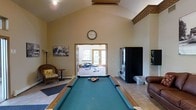 This single-floor recreational space is thoughtfully designed to serve as a hub for both leisure and fitness activities. The main area is anchored by a full-size pool table with a teal felt surface, positioned beneath a soaring vaulted ceiling with exposed beam detailing that adds architectural character and a sense of openness to the room. Warm neutral-toned walls and tile flooring contribute to the space's clean, welcoming ambiance. Comfortable seating, including a cushioned wicker chair and a leather chair, are positioned along the perimeter, offering spots to relax between activities. A wall clock and framed artwork add a homey, lived-in feel to the room. A black vending machine is conveniently located along one wall, adding to the facility's practical amenities. Through a wide doorway, a dedicated fitness area is visible, equipped with exercise equipment and bathed in natural light streaming through large glass-panel doors that lead to the outdoors. The flow between the billiards lounge and the fitness room creates a cohesive multi-use environment. Overall, this space strikes a balance between casual recreation and active wellness, making it well-suited as a shared amenity facility.