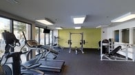 This fitness center is thoughtfully designed to support a full range of workout routines within a clean, modern environment. The space is laid out across a single floor and benefits from large windows along one wall, flooding the interior with natural light and providing views of the outdoors — creating an open, energizing atmosphere ideal for exercise. The flooring throughout is a durable rubber gym surface in a dark speckled finish, providing both safety and comfort underfoot. Overhead, large recessed panel lights ensure consistent, bright illumination throughout the space. The equipment selection is comprehensive: cardio machines including Star Trac treadmills and an elliptical trainer are positioned near the windows, while a power rack with a loaded barbell and an accompanying bench serve as the centerpiece for strength training. Additional cable and resistance machines are positioned along the perimeter, offering a well-rounded workout experience. An accent wall painted in a bold yellow-green adds a dynamic visual contrast to the otherwise neutral palette of whites and grays. A wall-mounted television is positioned for viewing during cardio sessions. The overall ambiance is functional yet inviting, making it a practical and motivating facility for daily fitness use.