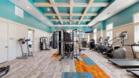 Located on Beach Boulevard in Jacksonville, Florida, this fitness center offers a thoughtfully designed single-floor space that balances energetic aesthetics with functional layout. The room's most distinctive architectural feature is its elevated coffered ceiling, where whitewashed wooden beams intersect against bold teal panels, creating a visually dynamic overhead focal point. Recessed lighting throughout ensures consistent illumination across the entire floor.

The walls are painted in a vivid teal above a neutral white wainscoting, lending the space an energetic yet clean atmosphere. Large windows along multiple walls allow ample natural light to fill the room, complementing the bright interior palette. A full-length mirror on one wall visually expands the space and supports strength training activities.

The flooring features a combination of neutral modular carpet tiles accented by a bold geometric pattern in orange, teal, and gold, defining workout zones across the open floor plan. Equipment is diverse and well-distributed, including treadmills, ellipticals, stair climbers, multi-station cable machines, a heavy punching bag, selectorized strength machines, a dumbbell rack with bench, and cycling equipment. A wall-mounted television provides entertainment during workouts. Near the entrance, a storage unit and sanitizing station support user convenience.