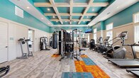 Located on Beach Boulevard in Jacksonville, Florida, this fitness center offers a thoughtfully designed single-floor space that balances energetic aesthetics with functional layout. The room's most distinctive architectural feature is its elevated coffered ceiling, where whitewashed wooden beams intersect against bold teal panels, creating a visually dynamic overhead focal point. Recessed lighting throughout ensures consistent illumination across the entire floor.

The walls are painted in a vivid teal above a neutral white wainscoting, lending the space an energetic yet clean atmosphere. Large windows along multiple walls allow ample natural light to fill the room, complementing the bright interior palette. A full-length mirror on one wall visually expands the space and supports strength training activities.

The flooring features a combination of neutral modular carpet tiles accented by a bold geometric pattern in orange, teal, and gold, defining workout zones across the open floor plan. Equipment is diverse and well-distributed, including treadmills, ellipticals, stair climbers, multi-station cable machines, a heavy punching bag, selectorized strength machines, a dumbbell rack with bench, and cycling equipment. A wall-mounted television provides entertainment during workouts. Near the entrance, a storage unit and sanitizing station support user convenience.