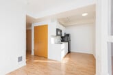 This thoughtfully designed single-floor space offers a clean, modern aesthetic throughout, making efficient use of its layout. The open living area is anchored by warm light wood flooring that runs seamlessly across the space, complemented by crisp white walls that enhance the sense of openness. A large window floods the room with natural light while framing views of the surrounding urban skyline.

The kitchen is neatly integrated into the living space via a compact peninsula layout. It features white cabinetry with sleek hardware, light-toned countertops, a stainless steel undermount sink, a black glass cooktop, an over-the-range microwave, and a full-size refrigerator — all finished in a cohesive black-and-white palette that gives the kitchen a contemporary feel.

The bathroom is well-appointed with a white undermount vanity sink, chrome fixtures, and a large mirror. A glass-enclosed shower adds a polished touch. Notably, the space incorporates an in-unit stacked washer and dryer, tucked efficiently beside the bathroom — a highly practical amenity that adds everyday convenience.

Natural wood-toned doors provide warm contrast against the predominantly white interior. Track lighting and recessed fixtures ensure the space remains well-lit throughout. Overall, the unit presents a functional and modern living environment with a clean, minimalist character.