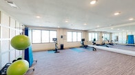 Located at 6490 Iron Horse Blvd in North Richland Hills, TX, this dedicated yoga and exercise room offers a well-equipped, open-plan fitness space designed to support a variety of workout routines. The room is finished with striped low-pile carpeting throughout and features recessed lighting that provides consistent, even illumination across the entire floor.

Large windows along the exterior walls allow natural light to fill the space while offering open views of the surrounding landscape, creating a bright and welcoming workout environment. A full-length mirrored wall with an attached barre runs along one side of the room, making it well-suited for yoga, stretching, barre workouts, and form-focused training.

Equipment in the room includes a stationary spin bike, a rowing machine, and a rack of dumbbells in graduated sizes. A freestanding equipment rack holds an assortment of stability and medicine balls alongside a reference chart for guided exercises. Yoga mats are available for floor-based training, and rubber flooring mats are placed beneath cardio equipment to protect the surface.

The walls feature a structured paneled design that adds a clean, polished aesthetic to the room. Informational signage is posted near the entrance. The overall layout provides ample open floor space, making the room versatile for group classes, individual workouts, or mindfulness practices.