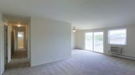 This single-floor space offers a clean, open layout defined by neutral walls and soft, plush wall-to-wall carpeting that adds warmth underfoot. The living area is generously proportioned, providing ample room for flexible furniture arrangements. A ceiling fan with a built-in light fixture is centrally positioned, offering both air circulation and ambient lighting. Natural light flows into the space through a large sliding glass door, which opens onto a private balcony enclosed by a white railing—offering a pleasant outdoor connection and views of surrounding greenery. A baseboard heating unit runs along the exterior wall beneath the sliding door, ensuring year-round comfort. The walls are smooth and painted in a soft, neutral tone, creating a bright and airy atmosphere throughout. Electrical outlets are thoughtfully distributed along the walls to accommodate a variety of room configurations. Overall, the space maintains a simple, functional aesthetic with a light and welcoming ambiance well-suited for comfortable everyday living.