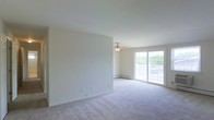 This single-floor space offers a clean, open layout defined by neutral walls and soft, plush wall-to-wall carpeting that adds warmth underfoot. The living area is generously proportioned, providing ample room for flexible furniture arrangements. A ceiling fan with a built-in light fixture is centrally positioned, offering both air circulation and ambient lighting. Natural light flows into the space through a large sliding glass door, which opens onto a private balcony enclosed by a white railing—offering a pleasant outdoor connection and views of surrounding greenery. A baseboard heating unit runs along the exterior wall beneath the sliding door, ensuring year-round comfort. The walls are smooth and painted in a soft, neutral tone, creating a bright and airy atmosphere throughout. Electrical outlets are thoughtfully distributed along the walls to accommodate a variety of room configurations. Overall, the space maintains a simple, functional aesthetic with a light and welcoming ambiance well-suited for comfortable everyday living.