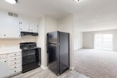 This single-floor space is thoughtfully laid out to maximize functionality across its key living areas. The kitchen is equipped with black appliances, including a coil-burner electric range with an overhead ventilation hood and a full-size top-freezer refrigerator. White cabinetry with black hardware lines the walls above and below the countertops, providing ample storage space. Wood-look tile flooring grounds the kitchen area, creating a clean, practical surface.

Adjacent to the kitchen, the open living area transitions to plush wall-to-wall carpeting in a neutral tone, offering a comfortable and cohesive feel. A sliding glass door fills the living room with natural light and provides direct access to an outdoor space. The bedroom is generously proportioned, featuring carpeted flooring, a ceiling fan with an integrated light fixture, and a window that allows natural light to filter in. A walk-in closet with built-in shelving offers substantial storage capacity.

The bathroom includes a white vanity countertop with an undermount sink and dark-finish faucet fixtures, complemented by a large wall mirror and a tiled tub surround. Throughout the space, neutral walls and clean white trim contribute to a bright, understated ambiance.