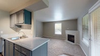 This single-floor residence offers a functional and comfortable layout across its living, kitchen, bedroom, and bathroom spaces. The open-concept living area is anchored by a tile-surround fireplace, creating a welcoming focal point. Soft carpet flooring extends throughout the living and bedroom areas, while wood-look vinyl flooring adds warmth and durability in the kitchen and bathroom. Natural light flows in through multiple windows fitted with blinds, offering outdoor views and a bright atmosphere. A ceiling fan in the dining area near the entry door enhances comfort and airflow. The kitchen is equipped with stainless steel appliances, including a range with an overhead ventilation hood and a refrigerator, complemented by gray cabinetry, light stone countertops, and a peninsula that connects the kitchen to the main living space. Teal upper cabinets add a distinctive design accent. The bathroom features a single-sink vanity with stone countertop, wood-look vinyl flooring, and a full tub-shower combination. The bedroom is generously sized, carpeted, and includes a window with natural light and a baseboard heater. The overall color palette of neutral grays and warm tones gives the space a clean, cohesive aesthetic throughout.