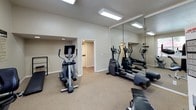 The River Terrace Fitness Center is a dedicated, single-floor exercise facility designed to support a well-rounded workout routine. The space is organized to accommodate both cardiovascular and strength training needs, with equipment distributed across an open, carpeted floor plan that promotes ease of movement and accessibility.

The cardio section is well-stocked, featuring multiple treadmills, recumbent and upright stationary bikes, and elliptical cross-trainers. A large wall-mounted mirror in the elliptical area enhances the sense of space and allows users to monitor their form during workouts. A multi-station cable and weight machine provides comprehensive strength training options alongside a dedicated workout bench.

Natural light flows into the facility through large windows with horizontal blinds, offering views of the surrounding greenery and creating a bright, welcoming atmosphere. Recessed fluorescent ceiling lighting ensures the space is well-lit throughout. The neutral beige walls and carpeted flooring contribute to a clean, comfortable environment.

Additional practical amenities include a wall-mounted sanitation station, a small waste bin, a security camera, and clearly marked exit signage, reflecting attention to cleanliness and safety. A posted fitness center rules and information board keeps users informed. The facility provides a convenient and functional setting for residents to maintain an active lifestyle.