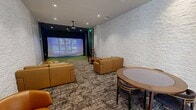 This contemporary amenity space combines a sophisticated lounge area with an immersive sports simulation experience, all within a generously proportioned single-floor layout. The lounge zone is anchored by a striking white stacked-stone accent wall adorned with large-scale artwork, creating a refined yet relaxed atmosphere. Warm cognac leather sofas and sectionals are arranged around natural wood slab coffee tables and a circular dining table with upholstered chairs, offering multiple seating configurations ideal for socializing or casual gatherings. Recessed ceiling lighting illuminates the space evenly, complemented by a patterned carpet that grounds the seating areas. A built-in wooden shelving unit with a media display adds a functional focal point. Floor-to-ceiling glass walls with black steel framing line one side, flooding the lounge with natural light and providing views to an adjacent space. At the far end, a dedicated sports simulator bay features vibrant green turf flooring, dark acoustic wall panels, dramatic navy draping, and a large projection screen capable of displaying golf, baseball, and other simulations. A ceiling-mounted projector serves both zones. Together, these elements create a dynamic, upscale environment well-suited for entertainment and recreation.