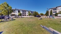 This contemporary apartment complex boasts a thoughtfully designed outdoor space that caters to both residents and their canine companions. The centerpiece is a generously sized dog park, enclosed by a chain-link fence for safety. The park's lush green grass provides a comfortable surface for dogs to play and exercise.

Within the dog park, several amenities enhance the experience for both pets and owners. A large blue shade sail offers respite from the sun, while benches positioned around the perimeter allow owners to relax and socialize. The area also includes agility equipment for dogs, such as ramps and obstacles, promoting active play and exercise.

The surrounding apartment buildings feature a modern architectural style with clean lines and a neutral color palette. Multi-story structures with balconies overlook the park, integrating indoor and outdoor living. Ample parking is available for residents, with both covered and open spaces visible.

Mature trees dot the landscape, providing natural shade and aesthetic appeal. The well-maintained grounds extend beyond the dog park, with manicured lawns and paved walkways connecting different areas of the complex. This thoughtful design creates a cohesive and inviting community space that prioritizes outdoor recreation and pet-friendly living.