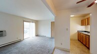 This single-floor residential unit offers a straightforward and functional layout designed for comfortable everyday living. The main living area is finished with neutral-toned wall-to-wall carpeting and features large sliding glass doors that allow natural light to fill the room while providing an outlook to the outdoors. A baseboard heating unit runs along the exterior wall, ensuring efficient climate control throughout the space. The crisp white walls and clean lines give the living area a bright, airy feel. A structural soffit detail adds subtle architectural definition to the ceiling, delineating the space without interrupting the open flow. Interior doors provide access to additional areas of the unit, including what appears to be a closet and a connecting hallway. The adjacent area, visible through an open passage, transitions to a lighter tile-style flooring, suggesting a secondary functional zone such as an entry, kitchen, or utility area. Overall, the space presents a clean, move-in ready environment with a simple, practical design well-suited for a variety of living arrangements.