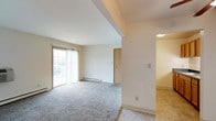 This single-floor residential unit offers a straightforward and functional layout designed for comfortable everyday living. The main living area is finished with neutral-toned wall-to-wall carpeting and features large sliding glass doors that allow natural light to fill the room while providing an outlook to the outdoors. A baseboard heating unit runs along the exterior wall, ensuring efficient climate control throughout the space. The crisp white walls and clean lines give the living area a bright, airy feel. A structural soffit detail adds subtle architectural definition to the ceiling, delineating the space without interrupting the open flow. Interior doors provide access to additional areas of the unit, including what appears to be a closet and a connecting hallway. The adjacent area, visible through an open passage, transitions to a lighter tile-style flooring, suggesting a secondary functional zone such as an entry, kitchen, or utility area. Overall, the space presents a clean, move-in ready environment with a simple, practical design well-suited for a variety of living arrangements.