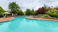 Located at 6115 Abbotts Bridge Rd in Johns Creek, Georgia, this outdoor amenity area showcases a generously sized freeform swimming pool as its central feature. The pool's curving, resort-inspired shape is complemented by crystal-clear turquoise water and a broad surrounding deck finished in warm terracotta-toned pavers, providing ample space for lounging and entertaining.

Numerous adjustable chaise lounge chairs are arranged along the pool deck, offering residents comfortable spaces to relax and take in the surroundings. A shaded outdoor seating area with a patio umbrella provides an additional gathering spot for those seeking respite from the sun.

The space is framed by mature, lush landscaping including tall deciduous trees, ornamental red-leafed shrubs, trimmed hedgerows, and natural stone accents, creating a private, park-like atmosphere. A wrought iron fence lines one perimeter, adding a classic architectural detail while maintaining an open, airy feel.

The backdrop features a well-appointed clubhouse building with traditional architectural elements including dormer windows, a chimney, and an expansive glass-paneled sunroom, reinforcing the upscale character of the community. The combination of thoughtful landscaping, quality amenities, and refined design elements makes this outdoor pool area a standout feature of the Johns Creek community.