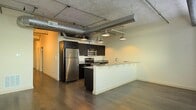 This single-floor space showcases a compelling industrial-modern aesthetic defined by exposed silver ductwork running across the concrete ceiling, lending an urban loft character to the open layout. The kitchen serves as the focal point, featuring dark espresso cabinetry that contrasts sharply with light granite countertops on both the main prep area and the extended breakfast bar. Stainless steel appliances, including a full-size refrigerator, electric range, and built-in microwave, provide a sleek, contemporary finish. Pendant lights hang at varying heights from the ceiling, offering both functional task lighting and a stylish design element that complements the industrial aesthetic. Rich, dark hardwood flooring extends throughout the open-concept space, grounding the design and adding warmth against the neutral-toned walls. The generous open floor plan between the kitchen and living area allows for flexible furniture arrangements, while the overall design balances raw industrial elements with modern finishes to create a cohesive and functional living environment.