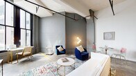 This single-floor loft masterfully balances industrial character with contemporary design. The open living area commands attention with soaring ceilings, exposed brick walls painted in a cool grey hue, and visible structural beams and piping that celebrate the building's raw heritage. Polished concrete floors run throughout, grounding the space with an urban authenticity.

The living area is anchored by a vibrant, multicolored area rug layered beneath a pair of plush navy velvet armchairs and a sleek round marble-top coffee table. A sculptural multi-globe floor lamp provides warm ambient lighting. Tall, dark-framed windows flood the space with natural light while offering city views, and a dedicated workspace with a desk and chair sits comfortably nearby.

The kitchen is outfitted with full stainless steel appliances, including a refrigerator, range, and dishwasher, complemented by crisp white cabinetry and a grey tile backsplash. A small dining area provides an intimate setting adjacent to the kitchen.

The bedroom features a striking gold four-poster canopy bed dressed in bold, botanically patterned bedding, paired with a matching arched gold-frame floor mirror. The bathroom offers a clean, functional layout with a shower-tub combination, mosaic tile accents, and white vanity cabinetry. Together, these spaces create a cohesive, design-forward loft with undeniable personality.