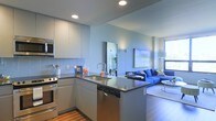 This single-floor space showcases a thoughtfully designed open-concept layout that seamlessly connects the kitchen and living area. The kitchen is outfitted with sleek, light gray cabinetry extending to the ceiling, providing generous storage. A central island with a dark countertop serves as both a functional workspace and a natural divider between the kitchen and living areas. Stainless steel appliances, including a dishwasher, range, and built-in microwave, lend a polished, modern aesthetic. A contemporary arc-neck faucet accents the undermount sink, while a classic white tile backsplash adds a clean, understated detail.

Recessed lighting throughout the kitchen ensures well-distributed illumination, complementing the natural light that enters through a large window in the adjacent living area. The living space is anchored by a bold blue sectional sofa and accented by vibrant wall art, creating a lively contrast against the cool, neutral-toned walls. A large mirror in the transitional space enhances the sense of depth and openness. Hardwood flooring ties the areas together, contributing warmth to the otherwise contemporary palette.