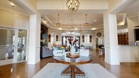 Located at 1709 Cason Ln in Murfreesboro, TN, this elegantly designed single-floor space offers a seamless open-concept layout encompassing a living room, dining area, and kitchen. Polished stained concrete floors flow throughout, complemented by soaring tray ceilings adorned with crown molding and an array of stylish pendant and chandelier lighting fixtures. The welcoming entryway features a glass-paneled door, wainscoting, and generous natural light. The expansive living area centers around a dramatic floor-to-ceiling stacked stone fireplace, paired with warm leather seating, cowhide accents, and arched mirrors that enhance the sense of depth. Large glass entry doors flood the space with abundant natural light and provide views to the outdoors. The kitchen is a standout, boasting dark espresso cabinetry, granite countertops, stainless steel appliances, a stone-accented center island with bar seating, and copper pendant lighting. Decorative columns frame the kitchen and common areas, adding architectural distinction. The dining area accommodates generous gatherings beneath an ornate chandelier. Throughout, the design strikes a balance between rustic warmth and refined elegance, creating an inviting and functional communal atmosphere.