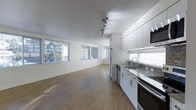 This single-floor space showcases a clean, contemporary design with an open-concept layout that seamlessly integrates the kitchen and living areas. The kitchen is outfitted with sleek white cabinetry, stainless steel appliances including a refrigerator, oven, and built-in microwave, as well as a stainless steel sink positioned beneath a window with a horizontal blind. The countertops and overall cabinetry design reflect a minimalist aesthetic, maintaining a bright and functional workspace.

The living area extends generously from the kitchen, offering an open and airy feel enhanced by multiple large windows along the far wall. These windows invite abundant natural light throughout the space while providing views of surrounding greenery and neighboring buildings. The warm hardwood-style flooring runs continuously throughout, adding a cohesive and welcoming element to the interior.

Track lighting mounted on the ceiling provides additional illumination and contributes to the modern character of the space. The white walls and smooth ceiling amplify the brightness and sense of openness. Overall, the space presents a practical yet stylish environment well-suited for comfortable everyday living.