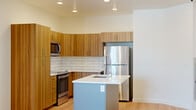 This thoughtfully designed single-floor unit offers a clean, modern aesthetic across a compact yet functional layout. The open-plan kitchen and living area form the heart of the space, with light hardwood flooring running throughout and recessed lighting illuminating the interior. The kitchen is a standout feature, showcasing warm wood-grain cabinetry that extends to the ceiling for ample storage, a white subway tile backsplash, and a full suite of stainless steel appliances including a refrigerator, range, microwave, and dishwasher. A central island with a crisp white quartz countertop and an undermount sink with a professional-style faucet provides additional prep space and serves as a natural divider between the kitchen and living area. The living area is bright and open, highlighted by large sliding glass doors that lead to a private balcony with views of the surrounding neighborhood, allowing abundant natural light to fill the room. The bathroom features a classic checkered tile floor in grey and white, a wood-accented vanity with a mirror, a toilet, and a full bathtub with shower. The overall palette of neutral whites, warm wood tones, and stainless steel finishes creates a cohesive, contemporary ambiance throughout the unit.