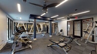 Located at 2202 State St in Nashville, Tennessee, this fitness center offers a well-appointed, single-floor workout facility designed with both function and aesthetics in mind. The space features a dark, sophisticated color palette with navy blue structural columns and ceiling accents contrasting against crisp white walls. Durable, patterned carpet tile flooring covers the workout area, providing both comfort and traction during exercise.

The facility is equipped with a comprehensive selection of strength training machines, free weights including a full dumbbell rack, adjustable benches, and cable machines, offering options for a complete strength workout. A dedicated cardio zone includes treadmills, an elliptical, and a stationary bike. Large floor-to-ceiling mirrors line one wall, visually expanding the space and supporting proper form during exercise. Decorative diagonal white trim frames the mirrors, adding a distinctive architectural touch.

Overhead, modern linear LED lighting runs across the ceiling, providing bright, even illumination throughout. Ceiling fans are strategically placed to maintain comfortable airflow. Glass panel doors along one wall open directly to an outdoor courtyard, introducing natural light and fresh air into the space. The overall ambiance is clean, modern, and energizing — well-suited for a focused fitness experience in the heart of Nashville.