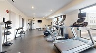 Located at 130 Fifth St SW in Rochester, Minnesota, this fitness center offers a well-appointed exercise room designed to support a comprehensive workout experience. The space features durable speckled rubber flooring throughout, providing both safety and comfort underfoot. Recessed lighting illuminates the room evenly, complementing the crisp white walls that give the space a clean, modern aesthetic.

The exercise room is thoughtfully equipped with a varied selection of cardio machines, including treadmills, elliptical trainers, and a stationary bike, positioned to make efficient use of the available floor space. A multi-function cable machine anchors the strength training area, accompanied by a weight bench and a dumbbell rack with a wall-mounted mirror for form checks. A pull-up and dip station rounds out the strength equipment offerings. A wall-mounted flat-screen television adds an entertainment element to enhance the workout environment.

Natural light filters in through a window along one wall, contributing to an open and energizing atmosphere. Acoustic panels on the walls help manage sound levels within the room. The space also includes a door providing direct access, making entry and exit convenient. Overall, this fitness center delivers a practical and well-organized environment suited for both cardio and strength training routines.