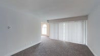 This single-story space offers a bright and open living area finished with wall-to-wall plush carpeting in a neutral tone, providing a comfortable and inviting foundation. The room is defined by a large floor-to-ceiling sliding door fitted with white vertical blinds, allowing abundant natural light to filter in while offering privacy and light control. A decorative arched window, positioned along the adjacent wall, adds an elegant architectural detail that complements the otherwise clean and minimalist interior. The smooth white ceilings and light-toned walls create a sense of openness and airiness throughout the space. The layout appears open and versatile, well-suited for a variety of furniture arrangements. White baseboards line the perimeter, adding a crisp, finished look to the room. Overall, the space presents a well-lit, move-in-ready environment with classic architectural touches and practical design elements that contribute to a comfortable and functional living experience.