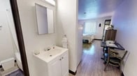 Located at 2500 University Blvd NE in Albuquerque, New Mexico, this efficiently designed one-bedroom, two-bathroom unit makes smart use of its compact footprint. The main living and sleeping area is the heart of the space, featuring warm wood-look flooring that runs throughout and crisp white walls accented by a striking bold orange feature wall that adds a vibrant, energetic character to the room. A window with vertical blinds allows natural light to filter in, brightening the interior. The bedroom area accommodates a full-sized bed and is complemented by a wall-mounted flat-screen TV, a multi-drawer dresser in a rustic wood finish, a dedicated desk and chair for work or study, open metal shelving for additional storage, and a couch that provides a casual seating area. A short entry corridor with a solid wood door leads into the unit, providing a clear separation from the outside. The space includes two bathrooms: one features a single vanity with cabinet storage and a framed mirror, while the second contains a shower-tub combination, toilet, and towel bar — both finished in clean, all-white palettes with textured walls. Light-tone wood-look flooring continues seamlessly through both bathrooms, unifying the overall aesthetic of the unit.