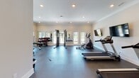 Located at 915 General Stewart Way in Hinesville, Georgia, this well-appointed fitness center offers a comprehensive single-floor workout facility designed to support a full range of exercise routines. The space features durable diamond-plate rubber flooring throughout, providing both safety and stability during workouts. Crown molding and recessed lighting lend a polished, finished look to the interior, while neutral walls and large windows allow ample natural light to fill the room.

The facility is equipped with a variety of cardio machines, including treadmills and an elliptical trainer, alongside a multi-station cable machine for strength training. A dedicated free weights section includes a fully stocked dumbbell rack with a variety of weight increments, an adjustable workout bench, and accessory equipment such as kettlebells and resistance tools. Full-length mirrors are strategically placed along the walls to support proper form during exercise.

A wall-mounted television provides entertainment during workouts. The space also includes a dual water fountain station for convenient hydration. Large windows and glass doors offer views of the surrounding outdoor pool area and community grounds. The overall layout is open and functional, offering an organized, clean environment well-suited for both cardio and strength-focused training sessions.