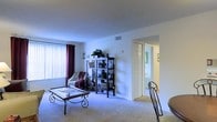 This single-floor residence features a living area finished with wall-to-wall carpeting that extends throughout the space, contributing to a warm and cohesive feel underfoot. The room is bathed in natural light from a large window dressed with vertical blinds and accented by deep burgundy curtains, providing both brightness and privacy control. Neutral white walls keep the space feeling open and versatile, while a ceiling-mounted HVAC vent and baseboard register indicate a comprehensive climate control system throughout.

A decorative open shelving unit serves as a focal point along the main wall, offering practical display and storage space. The living area accommodates a seating arrangement centered around a glass-top coffee table with ornate wrought-iron legs, adding a decorative touch to the room's functional layout.

An open doorway leads directly into an adjacent room, hinting at a connected and accessible floor plan that flows naturally between spaces. The bright, open passage suggests good interior lighting throughout. Overall, the space presents a clean, neutral palette with ample room for personalization, making it a practical and comfortable single-floor residence.