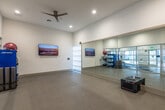 This single-floor fitness studio is designed with both functionality and visual energy in mind. The space is defined by its bold, geometric mural spanning multiple walls, featuring striking blocks of teal, yellow, red, and navy that create a dynamic and motivating atmosphere. Full-length mirrors line one side of the studio, enhancing the sense of openness and allowing users to monitor their form during exercise.

The workout area features a Life Fitness equipment rack stocked with kettlebells, medicine balls, and stability balls, alongside foam rollers and plyo boxes for a well-rounded training experience. A wall-mounted flat-screen TV is positioned for easy viewing during workouts. Recessed lighting throughout the ceiling provides bright, even illumination across the carpeted floor, which offers a comfortable surface for a variety of exercises.

Glass partition walls with aluminum framing separate the studio from an adjacent fitness area, maintaining visual connectivity while defining distinct zones for different activities. The glass walls also allow natural light to filter in, contributing to an airy, open feel. The clean white walls, high ceilings, and thoughtful layout make this studio a versatile and well-equipped space suitable for group classes, personal training, and individual fitness routines.